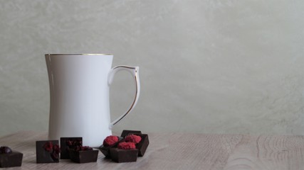 white mug and dark chocolate candies decorated with dried fruits on a wooden table on a light background
