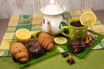 On the table is a cup of tea with lemon, a teapot. On the saucers are cookies, croissants and cinnamon sticks.