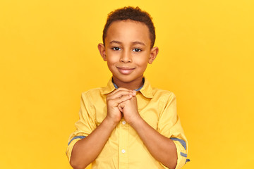 Studio shot of handsome adorable Afro American boy wearing casual yellow shirt clasping his hands...