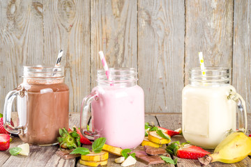 Three mason jars with milkshakes or smoothie. Summer healthy breakfast, lunch drinks - banana, chocolate and strawberry milkshakes on wooden background