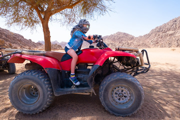 Fototapeta na wymiar Brave kid in checkered scarf around head is riding red quad bike in sand desert