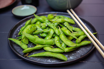 Cooked green Edamame lie on a dark plate in a Japanese restaurant.