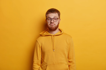 Horizontal shot of doubtful bearded man with ginger hair and beard purses lips and looks confusingly at camera, hears puzzled news, has specific appearance, wears yellow hoodie and spectacles