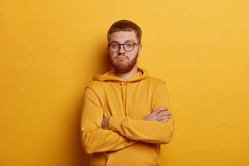 Fototapeta premium Self assured man with ginger hair and stubble, crosses arms over chest, being confident and boasts about his achievements, dressed casually, poses in yellow studio, doesnt trust friend looks at camera