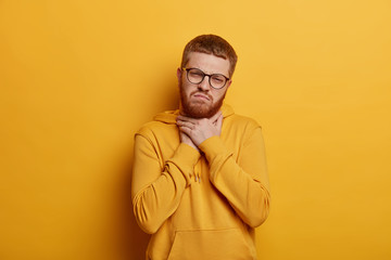 Upset young man has short ginger hair and bristle, touches neck and suffers from sore throat, has painful feeling while swallowing, wears hoodie, isolated over yellow background. Bad symptom