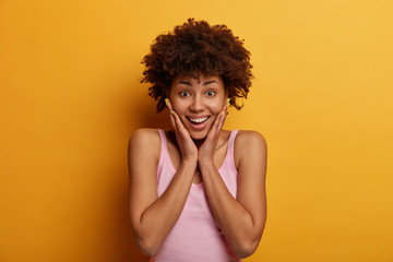 Positive emotions concept. Glad joyful curly African American woman touches cheeks, learned something unexpected and awesome, looks with happy smile at camera, poses over yellow studio background