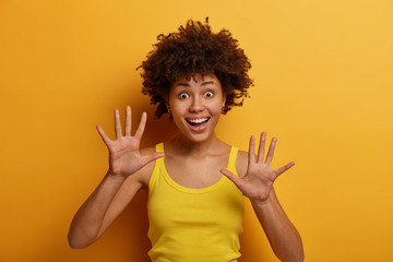 Positive dark skinned woman raises palms, feels joyful, has playful mood, gazes with funny expression, wears casual shirt, isolated over yellow background. People, emotions and happiness concept