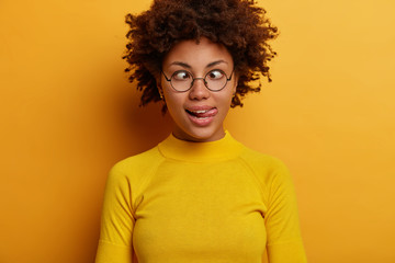 Childish funny woman with Afro hair sticks out tongue, crosses eyes, goes crazy and mad, makes grimace, wears round spectacles and casual jumper, poses against yellow background, has playful mood