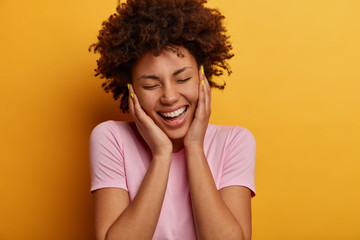 Fototapeta na wymiar Close up portrait of joyful curly haired woman touches cheeks, has sincere positive smile, closes eyes and being amused, laughs from something hilarious, dressed casually, isoalated over yellow wall