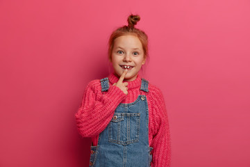 Little child with ginger hair knot points at two adult teeth, has glad expression, wears knitted sweater and denim sarafan, has positive mood, poses against rosy background. Childhood concept