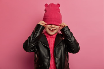 Positive small child hides face with pink hat, covers eyes, wears leather jacket, has playful happy smile, poses against rosy background, feels upbeat, tries on fashionable outfit. Children concept