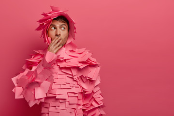 Wondered young man covers mouth, looks with frightened expression aside, covers with many sticky notes, wears creative paper made costume, isolated on pink background, blank space on right side