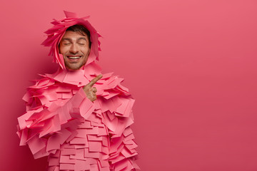 Delighted Caucasian man with funny positive expression indicates at blank space, advertises something with good mood, wears paper outfit made of adhesive notes, poses against rosy background