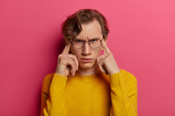 Photo of dissatisfied man suffers from headache, frowns face, touches temples with fingers, tries to focus, remembers something, wears spectacles, yellow sweater, isolated on pink background