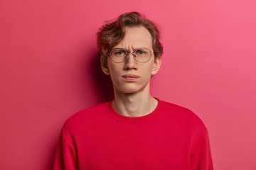 Horizontal shot of serious dissatisfied male model smirks face and looks straightly at camera, doubts he can trust you, wears spectacles and red sweater, isolated on pink background, feels intense