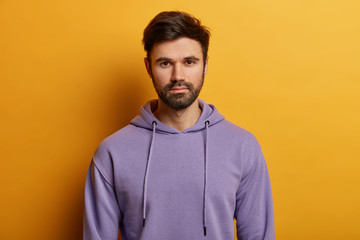 Serious handsome bearded European guy looks directly at camera, has thick bristle, wears purple hoodie, dressed in casual hoodie, poses over yellow background, listens information carefully.