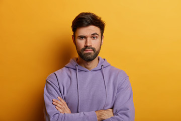Fototapeta premium Serious looking bearded adult man holds hands crossed, looks assertive at camera, raises eyebrows with wonder, judges presentation of colleague, wears hoodie, isolated over yellow background