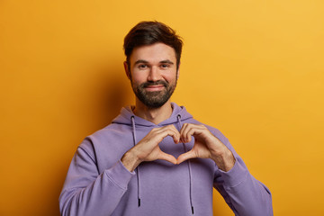 Friendly looking bearded guy shapes heart gesture, sends love, charity and volunteer, wears purple sweatshirt, poses over yellow background, demonstrates affection, makes valentine hand sign