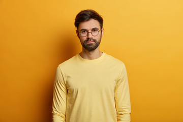 Photo of serious looking man has dark bristle, wears round spectacles and yellow jumper, direct gaze at camera, poses indoor, has casual talk with someone. Monochrome. Face expressions concept