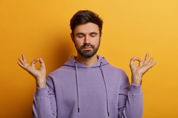 Fototapeta na wymiar Peaceful patient bearded man raises hands sideways with zen gesture, keeps eyes closed, rests after work or studying, being patient, poses against yellow background, breathes deeply and feels relieved