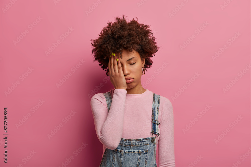 Wall mural fatigue curly haired woman feels bored and distressed, wants to sleep, covers half of face with palm