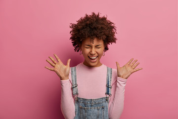 Playful positive young African American woman raises palms, goes crazy about hear amazing news, laughs out, dressed in turtleneck and sarafan, poses over pink background. Happy emotions concept
