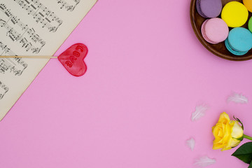 Pink background with antique music sheets, french macarons sweets, yellow rose, heart shaped lollipop love and small white soft feathers.Top view. For designs, concepts or posters.