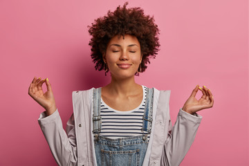 Portrait of relieved determined dark skinned woman meditates with closed eyes, practices breathing exercise or yoga to calm down, stands in lotus pose, poses over pink background, makes zen gesture © WHstudio Leushin N