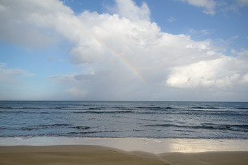 Rainbow, beautiful blue sky and fluffy clouds above the empty sandy beach. Natural Seascape.