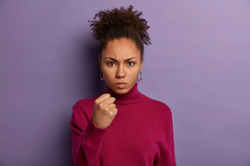 Strict young female teacher shows clenched fist at camera, discontent with pupils behaviour, promises to punish, looks angrily, has curly combed hair, wears burgundy jumper, isolated on purple wall