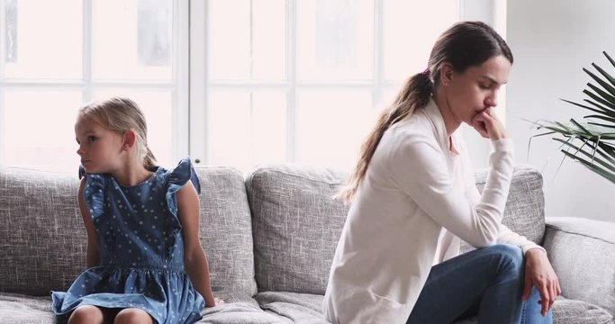 Unhappy mother and child daughter sit on sofa avoiding talk
