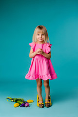 little blonde girl in big yellow shoes and a pink dress is holding spring flowers, baby girl in a pink dress and mother's shoes smiling on a blue background, a little fashionista. space for text