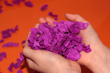 In the hands of small pieces of purple paper. Children's hands on an orange background. Fun game.