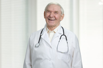 Old cheerful doctor is laughing. Male senior wrinkled physician with stethoscope. Windows with jalousies background.