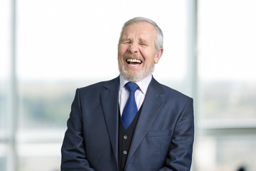 Old manager in office laughing hard. Cheerful boss in business suit portrait. Blurred windows background.