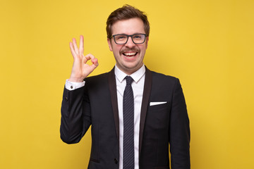 Happy young man with mustache in shirt and tie showing OK sign and smiling om yellow wall.