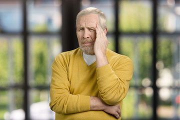 Portrait of old grandpa having headache. Touching and massaging head, feeling pain. Checkered windows background.