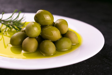  Green olives and olive oil with rosemary in a white plate on a black background.