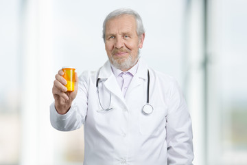 Old senior doctor giving orange can of pills to you. Bright blurred background.
