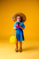 little blonde girl in a straw hat and sunglasses is smiling on a yellow background. child girl in a blue dress is holding a pineapple-shaped drink glass on a yellow background.summer vacation concept