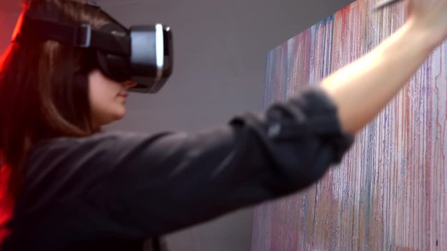 Talented female artist wearing new virtual reality glasses, using two paint brushes and creating emotional picture on large canvas in art studio. Interactive game with modern art and colours