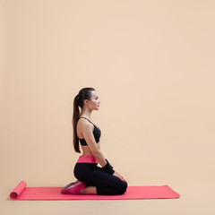 Fototapeta premium A young attractive girl with a ponytail, sits on knees on pink yoga mat, ready for classes, stretching, pink background.
