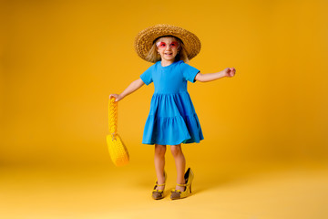 little blonde girl in a straw hat and sunglasses is smiling on a yellow background. child girl in a blue dress is holding a pineapple-shaped drink glass on a yellow background.summer vacation concept