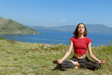 Naklejka na ściany i meble Young woman doing yoga and meditating in mountain landscape. Concentrated and contented female sitting with closed eyes in lotus position at grass with lake behind in sunny day 