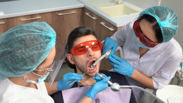 A professional dentist works with the help of an assistant. A male client of a clinic sits with his mouth open in a dental chair, wearing safety glasses. Dental office. Dentistry. Stomatology