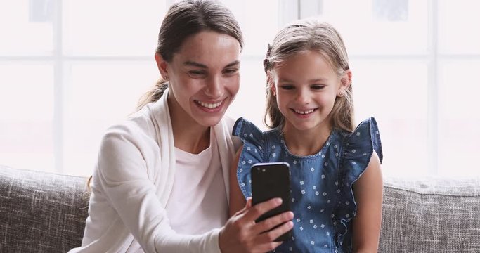 Cheerful mum showing cute small daughter funny video on smartphone