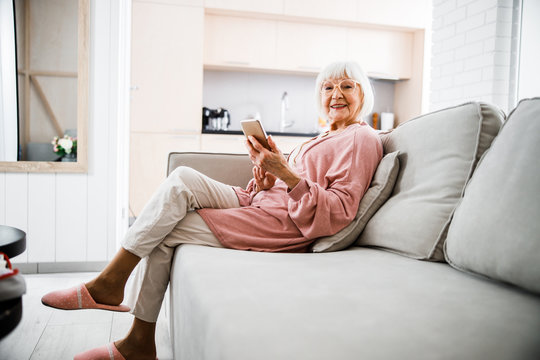 Smiling elderly woman sitting on couch and holding smartphone