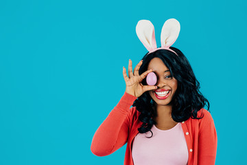 Portrait of a smiling happy young woman holding Easter egg over eye and bunny ears, isolated on...