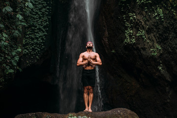 A man of athletic build does yoga. Healthy lifestyle. The concentration of the body. A man does yoga at a waterfall. A man does yoga in Bali. A man meditates in nature. Meditation at the waterfall