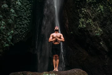 Wandaufkleber A man of athletic build does yoga. Healthy lifestyle. The concentration of the body. A man does yoga at a waterfall. A man does yoga in Bali. A man meditates in nature. Meditation at the waterfall © MISHA